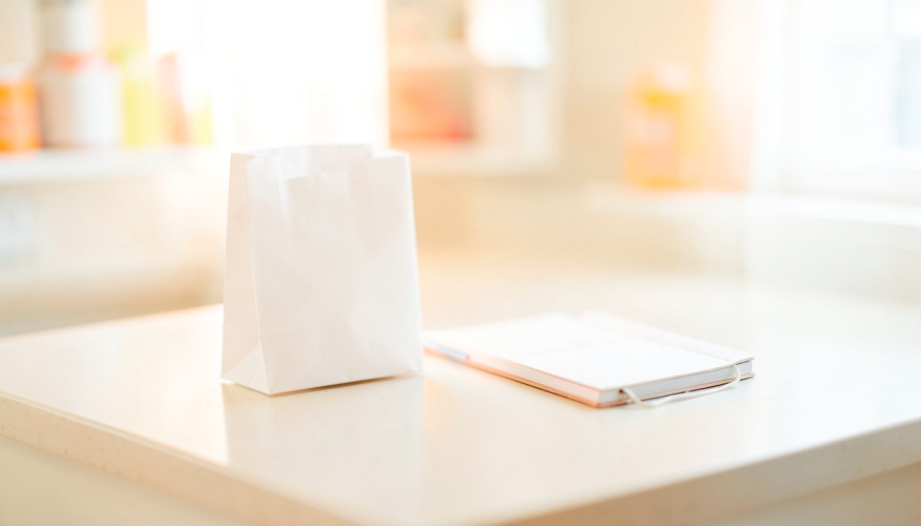 Soft-focus photograph of a clean pharmacy countertop with a small white paper bag and a notepad, bathed in warm natural light with cream and pale blue tones